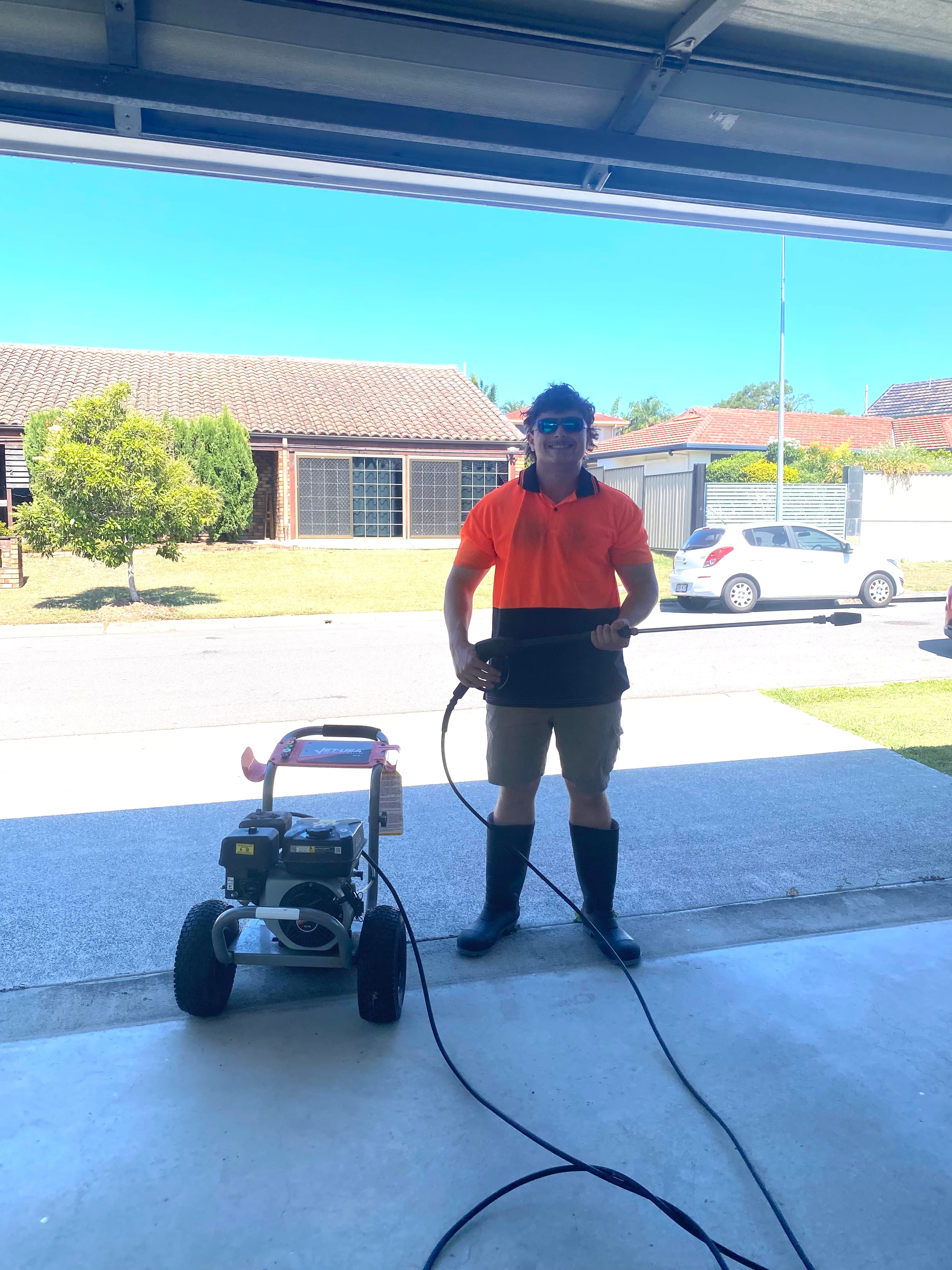 Man in high-visibility clothing holding a pressure washer wand in a sunny garage opening.